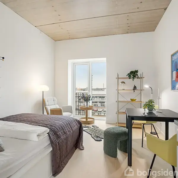 Værelse med seng og skrivebord, belyst af en gulvlampe. I baggrunden ses en balkondør og udsigt over bygninger. Væggene har minimal kunst, og rummet er lyst og moderne.