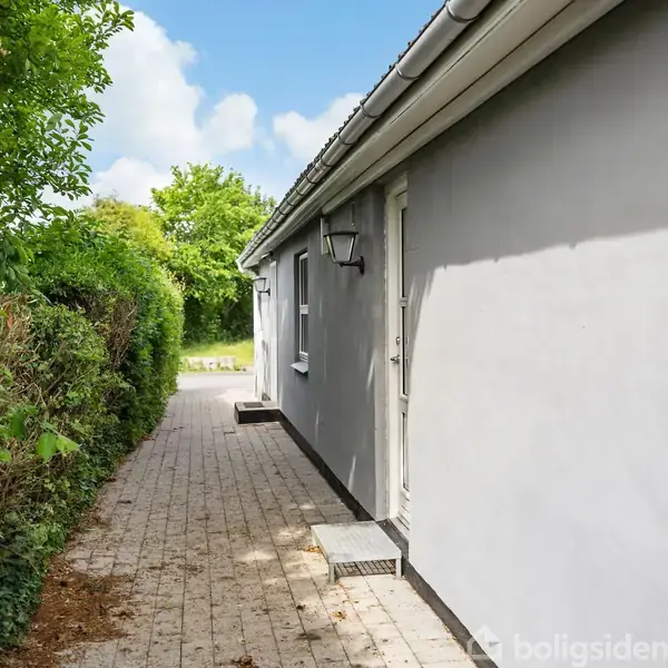 Hus med hvid facade i et haveområde. En flisebelagt sti løber langs muren, omkranset af grønne buske. En lampe hænger ved døren under den blå himmel.