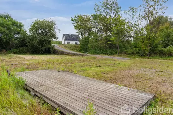 En træplatform ligger på en åben græsplæne. I baggrunden ses en hvid bygning med et sort tag omgivet af tætte grønne træer og en snoet grusvej.