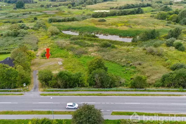 En rød pil peger mod en tom byggeplads omgivet af frodig natur med buske og små søer. I forgrunden kører en hvid bil på en vej.