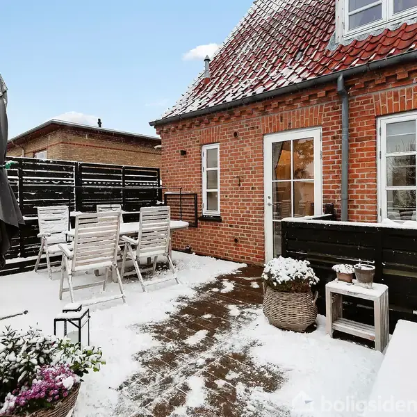 Terrassen har et snedækket træbord og stole, der er omkranset af mørke trælåger. Sollys skinner fra den kolde blå himmel, mens sne dekorerer taget på det røde murstenshus.