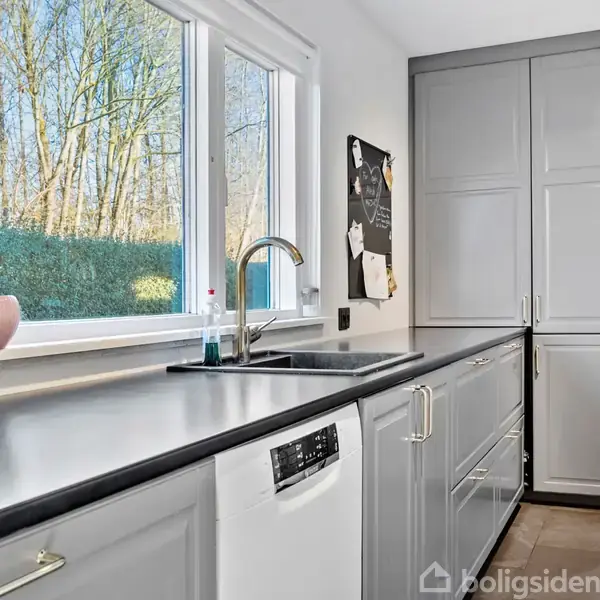 En køkkenbordplade med en elkedel står ved et vindue. Opvaskemaskinen er indbygget under bordet. Grå skabe omgiver området, set fra et rummeligt, lyst køkken.