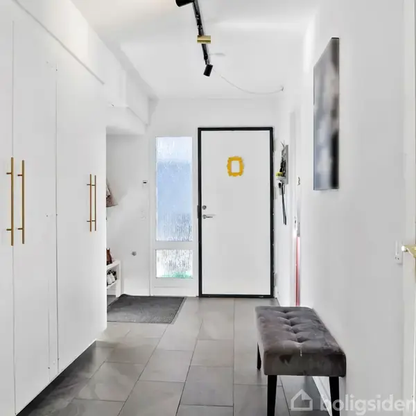 En korridor med hvide skabe til venstre, en mørk polstret bænk til højre, og en dør i baggrunden. Navnet "LUCY" er placeret på væggen ved siden af døren.