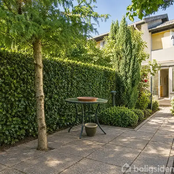 Rundt havebord står stille på flisebelagt terrasse omgivet af tæt klippet hæk og grønne planter. Baggrund viser et hus med store vinduer. Tekst: "AI visualisering".