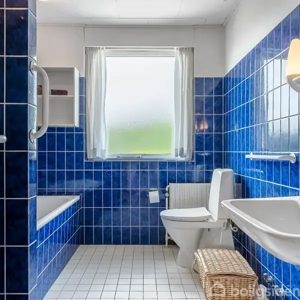 Badeværelse med blå fliser, et toilet og en håndvask. Et vindue med gardiner slipper lys ind. En kurv står ved siden af håndvasken. Der er ingen synlig tekst.