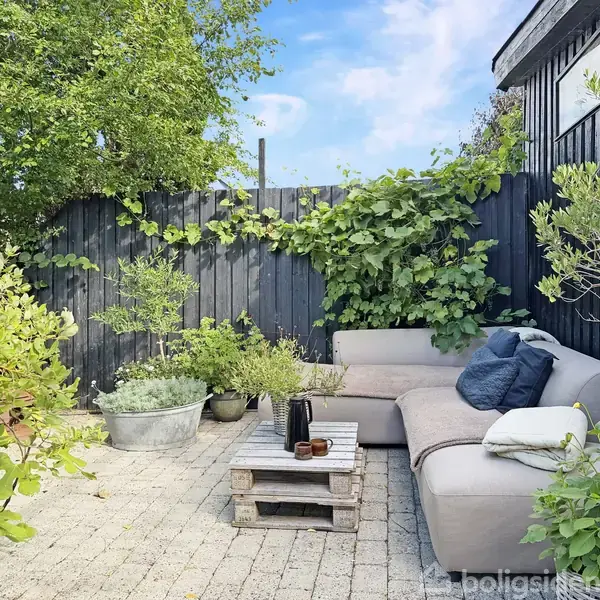 En beige udendørs sofa står på en terrasse, omgivet af grønne planter i potter foran et sort hegn. Et bord lavet af paller er placeret foran sofaen.