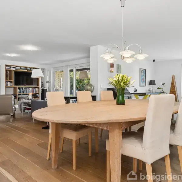 Et rundt spisebord i lyst træ omgivet af polstrede stole er centralt i rummet. Bordet er pyntet med en vase med blomster. I baggrunden ses en stue med bogreol og store vinduer.