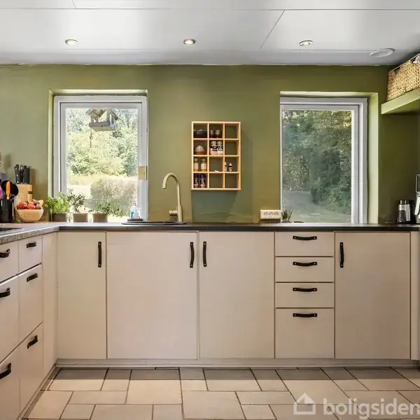 Et moderne køkken med hvide skabe, der indeholder køkkenudstyr. Køkkenet har vinduer med udsigt til grøn natur. Væggene er grønne, og der er en indbygget ovn til højre.