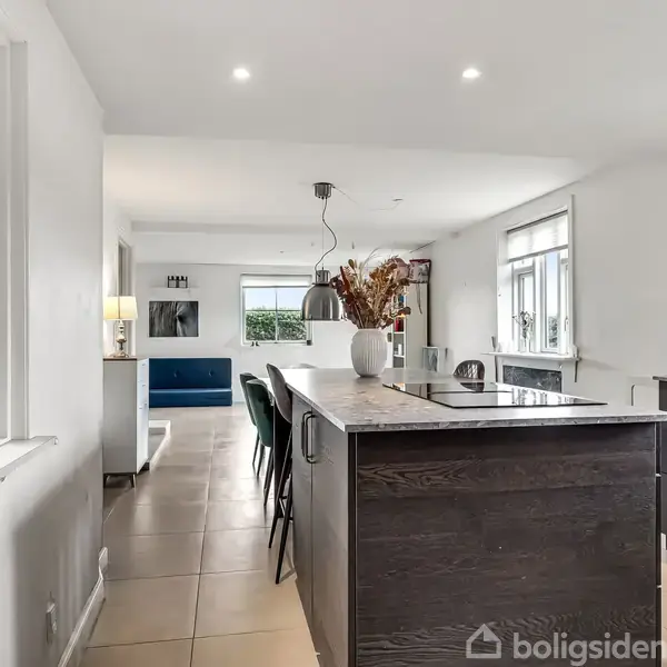 Køkken med mørk køkkenø fyldt med blomster, omkranset af barstole, i åbent lysegråt rum med vinduer, lyse fliser og spiseområde med sofa i baggrunden.