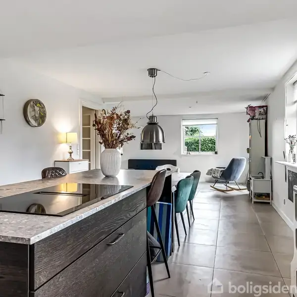 Køkkenø med kogeplade og vase med tørrede blomster; spisestole i flere farver omkring bord; stor lampe hænger over bordet; lyse vægge og vinduer giver rummelighed; i stuen bagved.