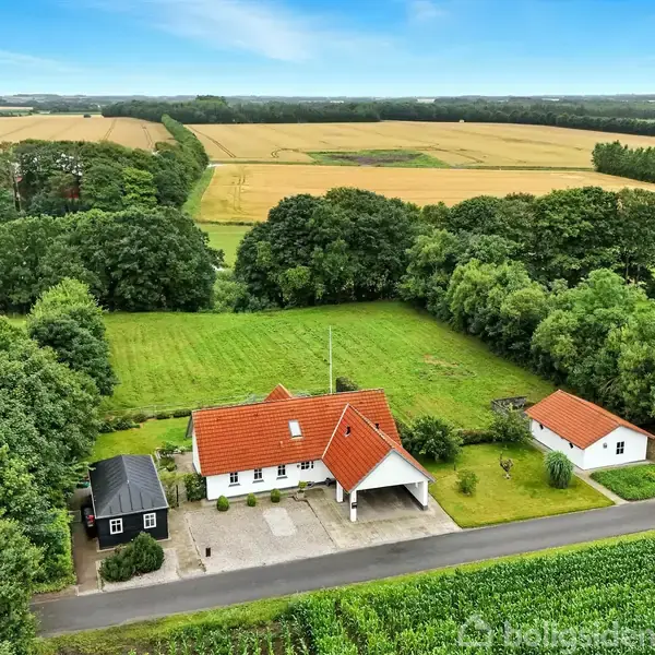 Et hus med rød tag ligger ved en landevej, omgivet af grønne marker og træer. Foran huset er en garage og indkørsel. To biler ses ved vejen.