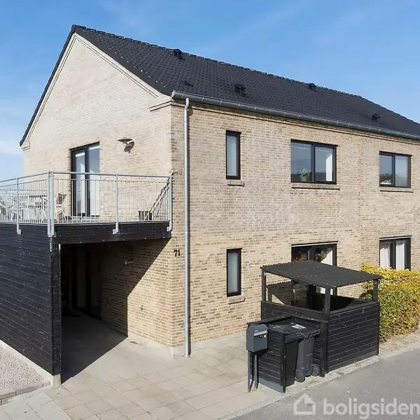 To-etagers murstenshus med sort tag ses fra gadesiden. En stor altan er tilknyttet, og forhaven omfatter hegn og skraldespande. Huset er beliggende i et boligkvarter.