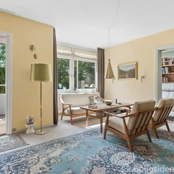 Stue med to lænestole og et sofabord på en mønstret blå tæppe. Store vinduer lader dagslys ind, og en lampe står i hjørnet. Udgang til altan med stole bagved. Bookshelf til højre.