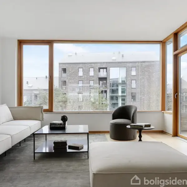 Stue med hvid sofa og lænestol, indrammet af store, lyse trærammevinduer, der giver udsigt til et lejlighedskompleks. Lavt sofabord med dekorative elementer står centralt på et gråt tæppe.