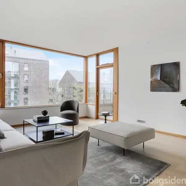 En lys stue med en stor hjørnesofa og sofabord foran store vinduer, der viser en udsigt over bygninger udenfor. En plante og kunstværk pryder rummet.