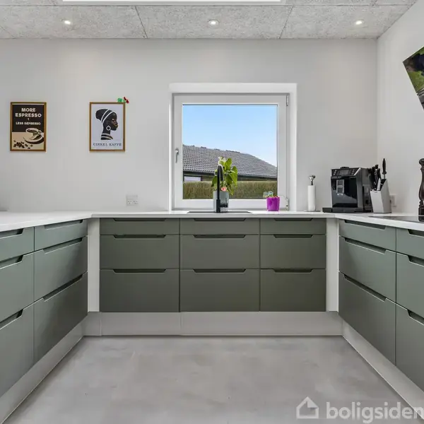 Køkken med grønne skabe, stilfulde køkkenmaskiner og planter på bordpladen. Vinduet i midten giver udsigt til en have. Væggen har kaffeposters: "Home Espresso" og "French Coffee".