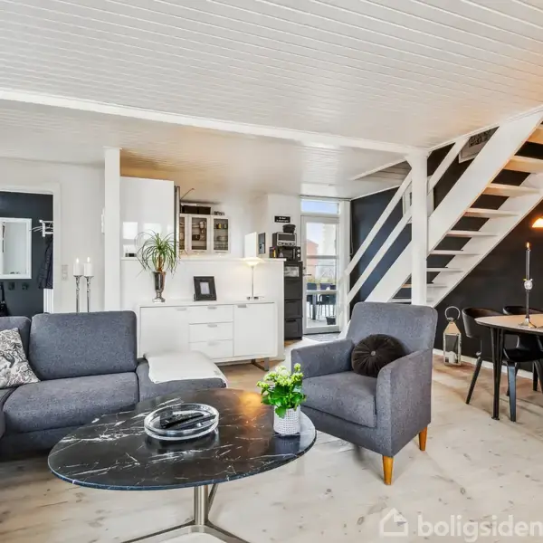 En moderne stue med en grå hjørnesofa og et rundt sofabord i midten. Ved siden af er der en spiseplads med et sort bord og stole. Baggrunden inkluderer en trappe til øverste etage.