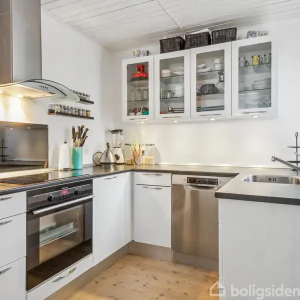 Et moderne, funktionelt køkken med hvide skabe og sorte bordplader. Skabe med glaslåger viser kopper og glas. Belysning ovenfor ovnen og overskabe. Krydderier og køkkenredskaber er synlige.