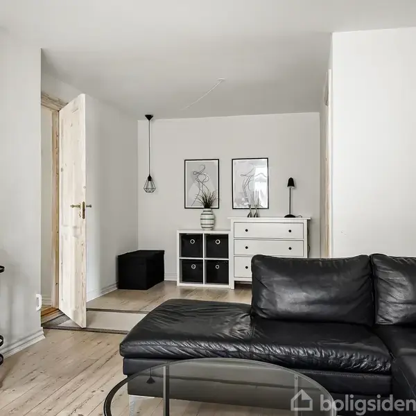 Sort lædersofa står i en lys stue med trægulv. To døre fører til andre rum. Hvide kommoder med sorte og hvide skuffer står under to indrammede billeder. En grøn plante pynter rummet.