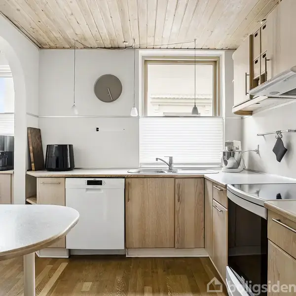 Køkken med lyse træskabe og hvide apparater, herunder opvaskemaskine og ovn. Rundt spisebord. Vindue med persienner og loftslamper. Trægulv og loft, ur på væggen.