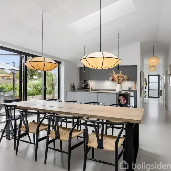 Spisebord omgivet af seks stole, placeret under to hængende lamper, i et lyst rum med åbne glasdøre mod en terrasse og have. Køkkenområde i baggrunden.