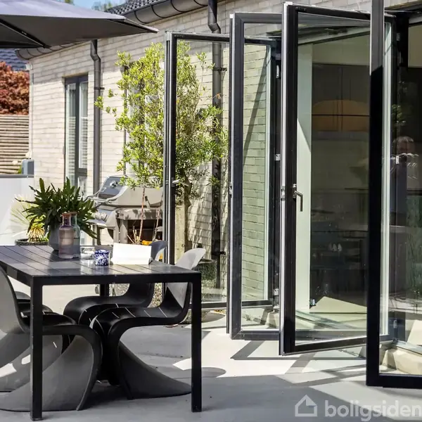 En moderne terrasse har et sort spisebord med kurvede stole. Bordet er placeret under en halvåben parasol, og terrassen er tilknyttet en bygning med store glasfoldedøre.