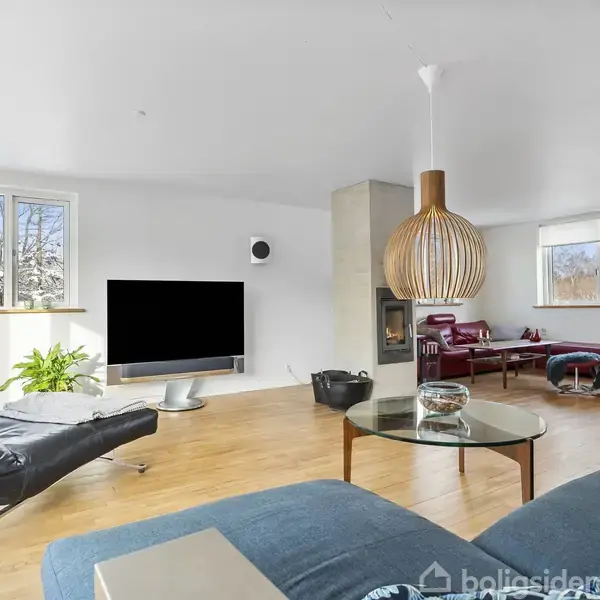 En moderne stue med en sort lænestol og fladskærms-TV, runde borde, og en stor trælampe. Store vinduer giver udsigt til et snedækket landskab udenfor.