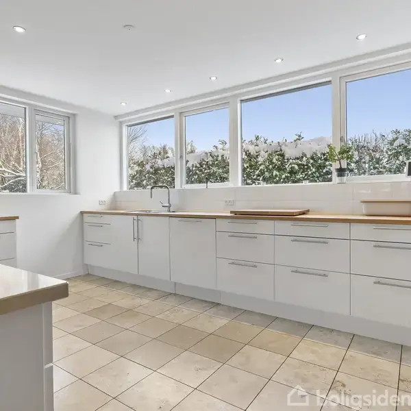 Et lyst køkken med hvide skabe og skuffer står tomt. Store vinduer i baggrunden giver udsigt til sneklædt have. Fliser dækker gulvet, og loftet har indbygget belysning.