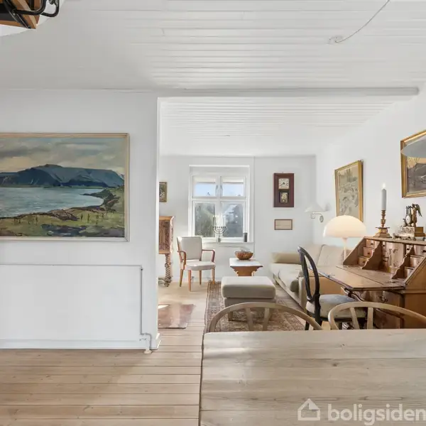 Stue med hvide vægge og trægulv, indeholdende et skrivebord med stol, sofaer, og malerier på væggene. Store vinduer slipper lys ind i rummet. Lampen hænger fra loftet.