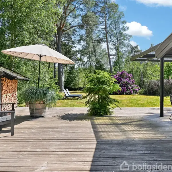 Terrasse med trædæk med parasol og havemøbler. Til venstre er der et skur fyldt med brænde. I baggrunden ses en græsplæne, blomsterbuske og store træer under en blå himmel.
