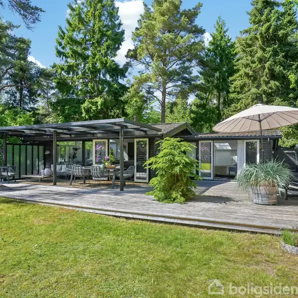Sommerhus med stor træterrasse, møbleret med stole og borde under parasol, omgivet af tæt skov. Terrasseområdet har opstablet brænde og dekorative planter i potter.