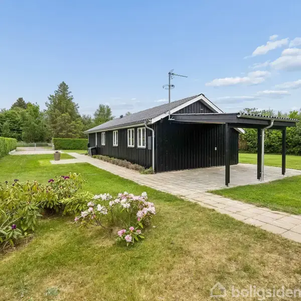 Sort hus med veranda står på en grøn græsplæne omgivet af buske. Flisesten leder fra verandaen gennem haven, mens klare himmelstræk omringer ejendommen.