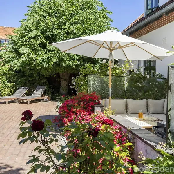 Parasol står på terrasse med lyse hynder og bord, omgivet af frodige buske og blomster. To liggestole er placeret på brostensbelægning ved siden af et stort træ i en velplejet have.
