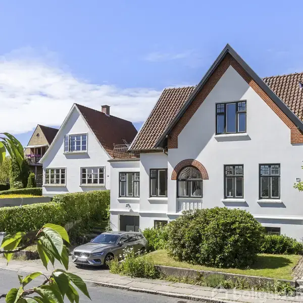Et hvidt hus med rødt tag står i en villakvarter. En bil er parkeret foran huset ved en stille vej, omgivet af grønne buske og træer under en blå himmel.