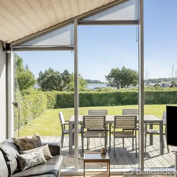 En stue med sort sofa i forgrunden vender mod store vinduer, der åbner ud til en terrasse med træbord og stole. Baggrunden viser en græsplæne og udsigt til vand og sejlbåde.