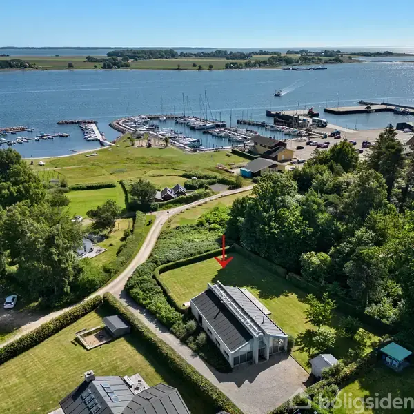Et hus ligger omgivet af frodig skov og græsplæner nær en marina. I baggrunden ses en kystlinje med bådepladser og havn, omgivet af åbent vand og små øer.