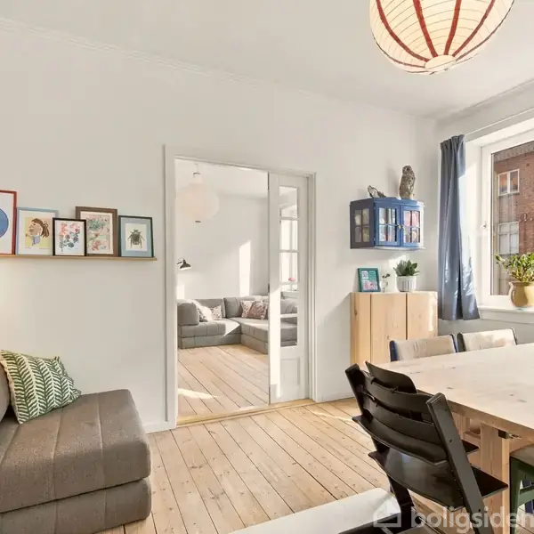 Stue med træbord og stole, sofa til venstre, kunst på væggen, åbning til tilstødende værelse, stor vindueskarm med planter, lysekrone, blå gardiner og udsigt til boligblok udenfor.