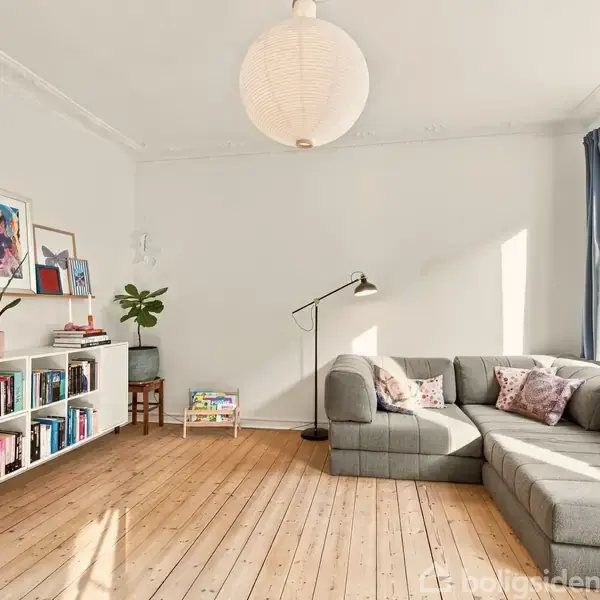Grå hjørnesofa står i en lys stue med trægulv og stor loftlampe. En bogreol langs væggen indeholder bøger og planter. Store vinduer lader sollys ind.