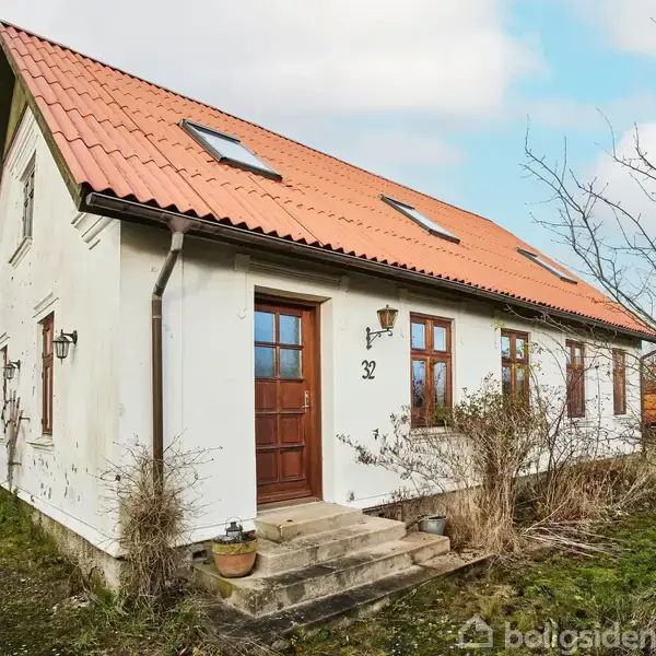 Et hvidt hus med rødt tag står i en have med sparsom beplantning. Huset har flere vinduer og en dør med tallet "32" ved indgangen.