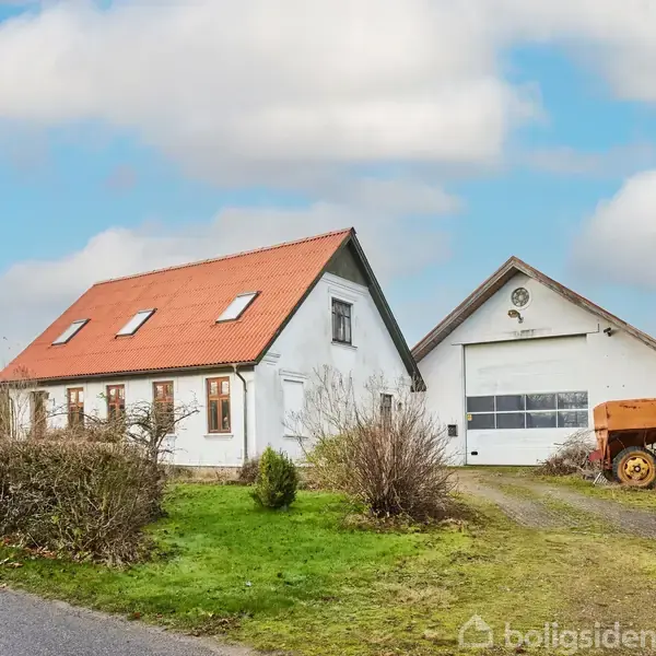 Et hvidt hus med rødt tag står ved siden af en stor garage med port. En gammel maskine er placeret ved garagen, og en vej løber langs den grønne have.