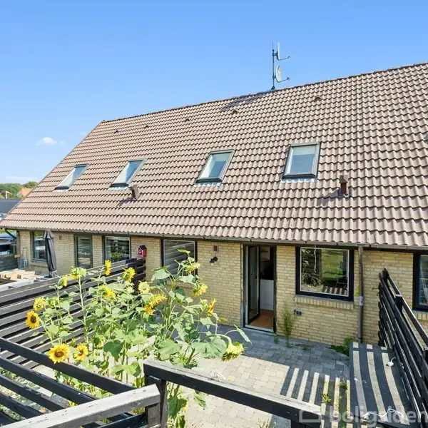 Rækkehus med rødt tegltag og flere vinduer, omkranset af en lille gårdhave med solsikker. Sort stakit afskærmer området. Solrigt vejr med blå himmel i baggrunden.