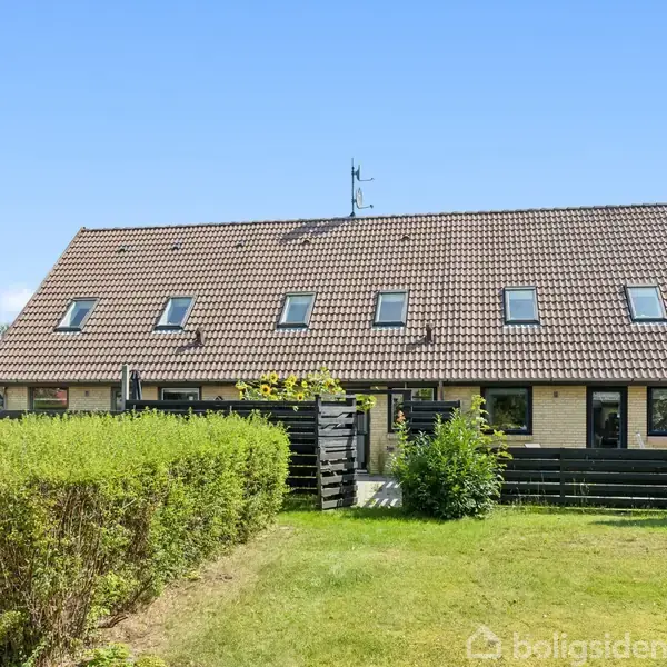 Et rækkehus med tegltag og flere kvistvinduer står i en grøn have omkranset af hække. Huset er placeret i et solrigt område med en blå himmel i baggrunden.