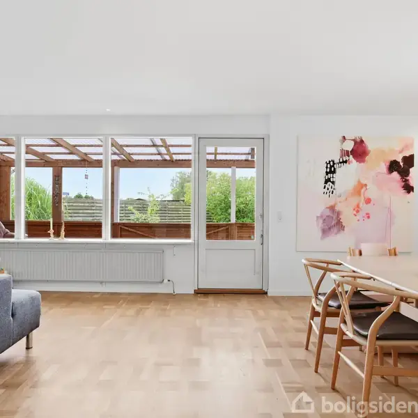 Stue med grå sofa og spisebord med stole, udsigt til terrasse gennem glasparti, lys maleri på væggen, træparketgulv og grøn udsigt udenfor.