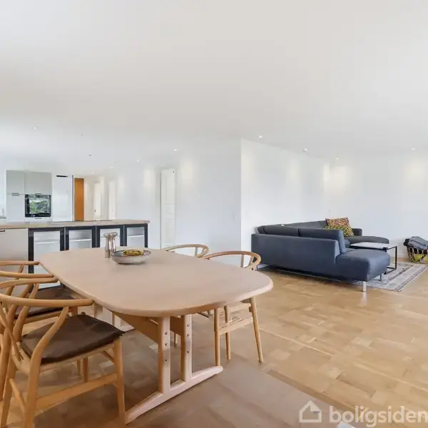 Et lyst og rummeligt stueområde med et spisebord og stole i forgrunden, en blå hjørnesofa til højre, og legetøjsruller i hjørnet. I baggrunden ses et køkkenområde.