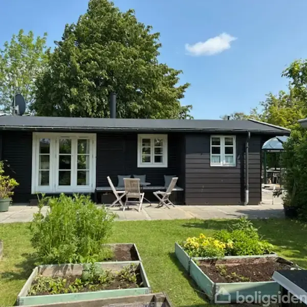 Et sort træhus står stille i en frodig have, omgivet af grønne planter og blomsterbede. Et par havemøbler står foran huset, mens himlen er blå og skyfri.
