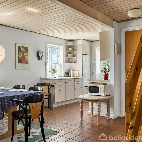 Køkken-alrum med spisebord dækket med blåternet dug, omgivet af sorte stole. Køkkenet i baggrunden har hvide skabe, mikroovn og køleskab. Trappen fører til øvre etage. Naturligt lys ind ad vinduer.