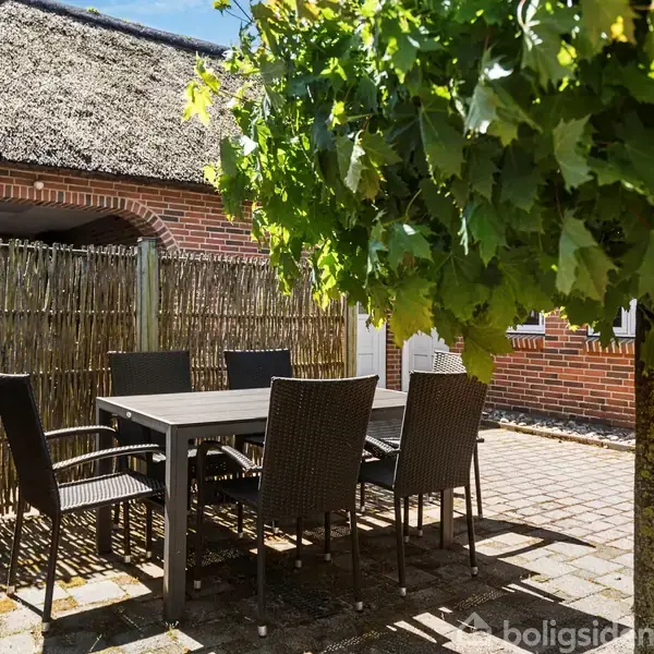 Et sæt udendørs møbler står på en flisebelagt terrasse. Bordet er omgivet af seks stole. En tæt busk giver skygge, og en stråtækt bygning ses i baggrunden.