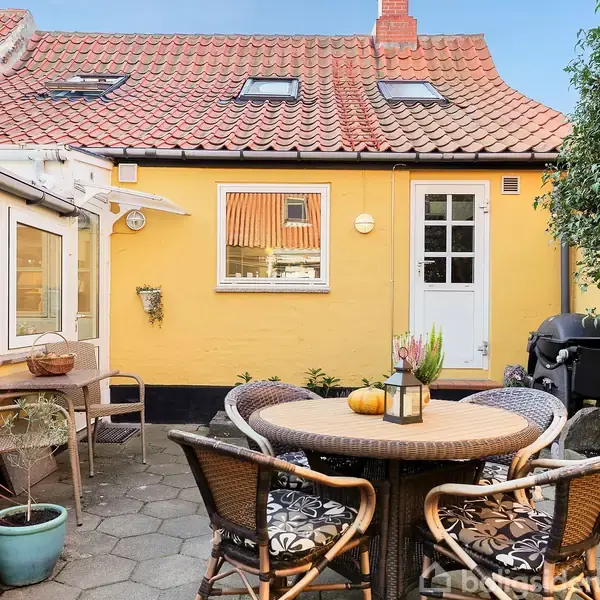 En rund terrassebordsgruppe med fire stole står på en stenbelagt gårdhave foran en gul murstenshusvæg. Der er blomsterkrukker og en grill i nærheden.