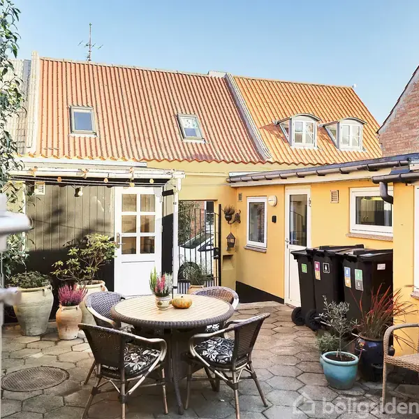 Et rundt bord med stole står i en brostensbelagt gårdhave, omgivet af planter i potter. Gule mure og et teglbelagt tag danner baggrunden med flere vinduer.
