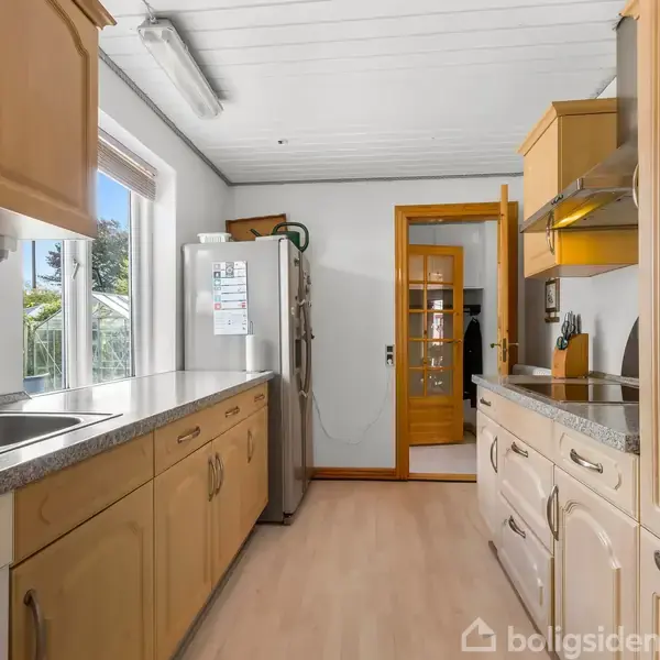 Køkken med lyse trækøkkenskabe, rustfri stål vask, og ovn. Sollys strømmer ind gennem et vindue til venstre. En metalkøleskab står ved en dør til et tilstødende rum.